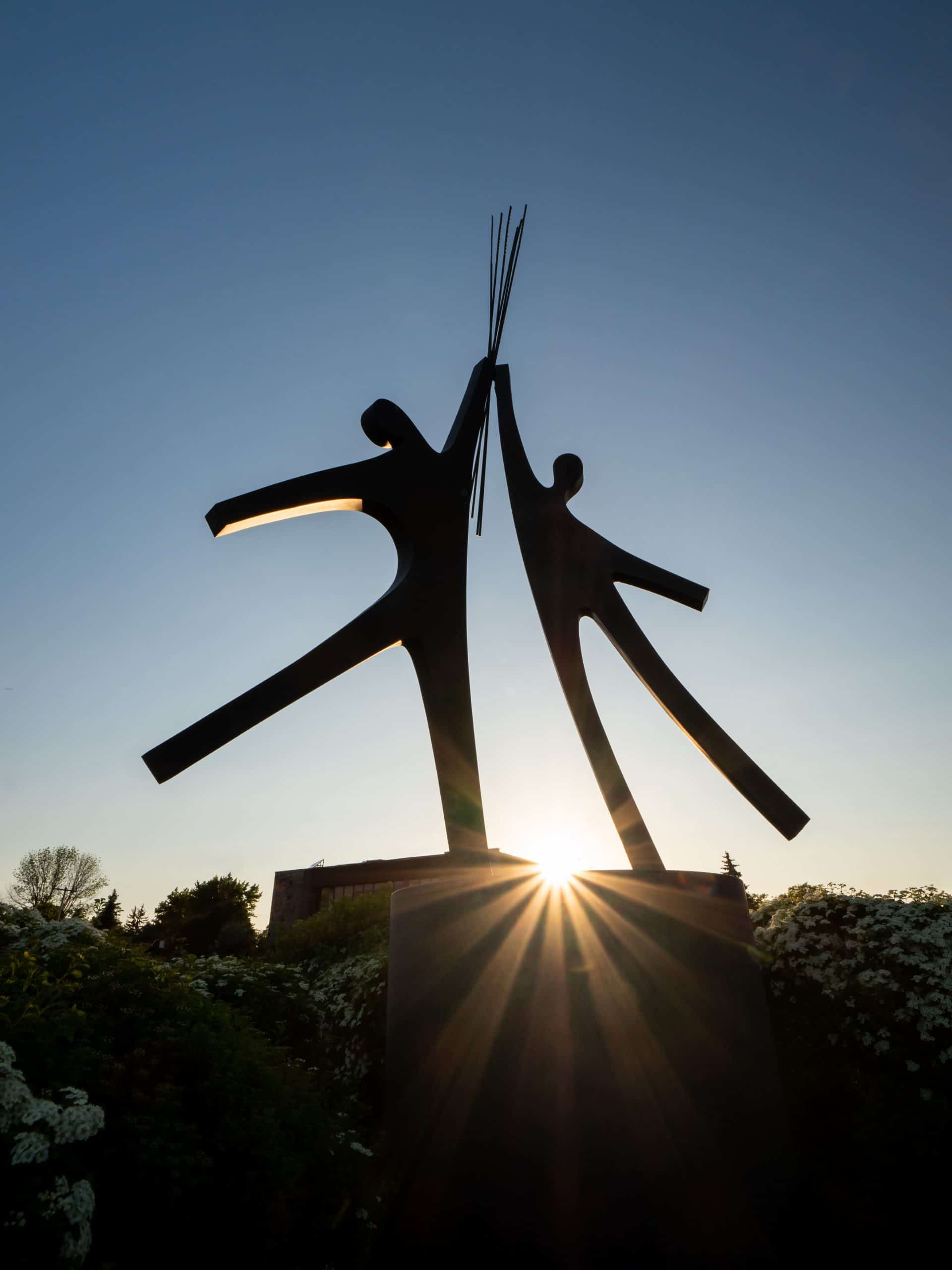 Catalogue d'art public - artiste Jacques Bénard - La Rencontre, sculpture en métal d'un couple tenant une gerbe de blé à bout de bras, vue de près. au coucher du soleil