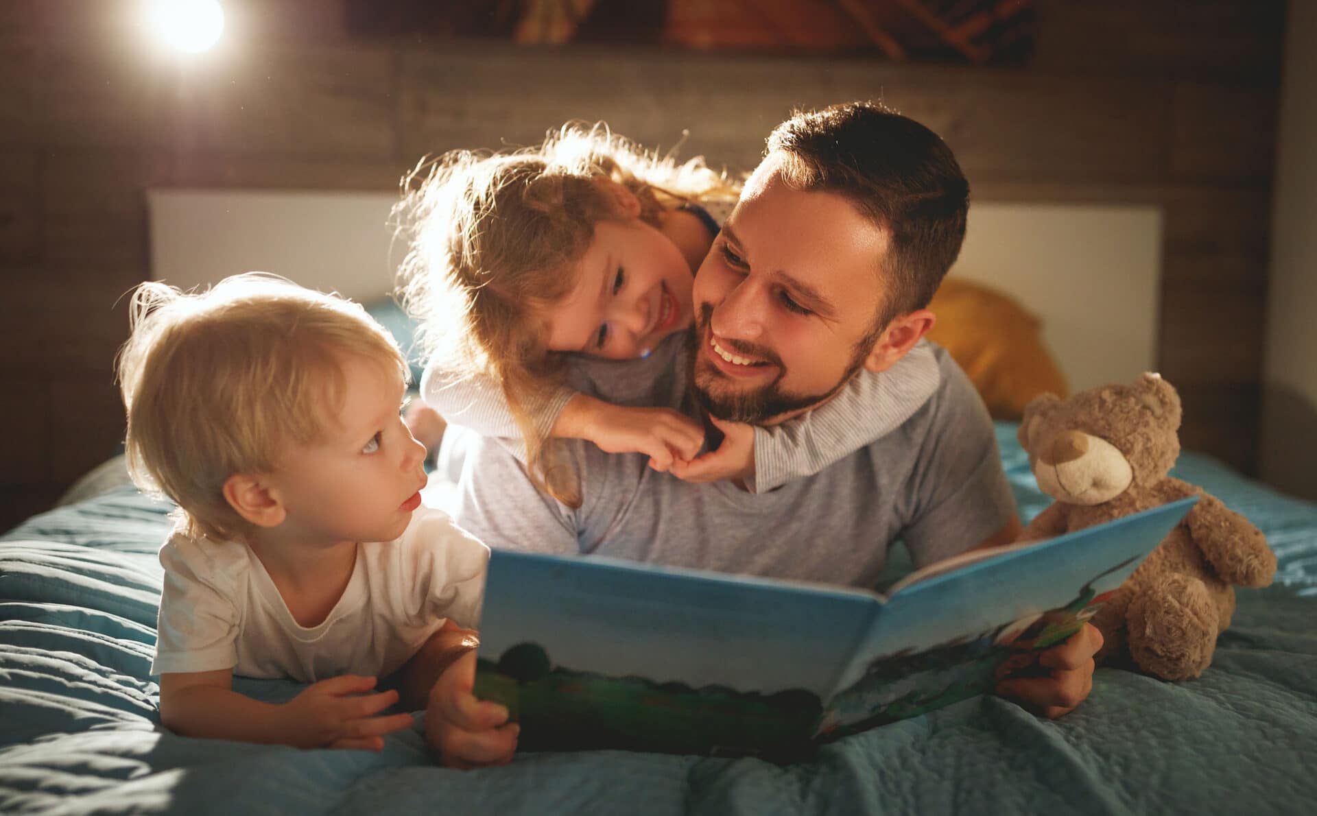 Un papa qui fait la lecture à ses deux jeunes enfants. Son petit garçon est assis à côté de lui et le regarde et sa petite fille est accrochée au cou de son papa et ils sourient.