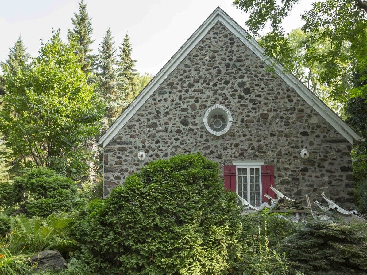 Maison en pierre avec des volets rouges à la fenêtre. Le tout entouré de verdure.