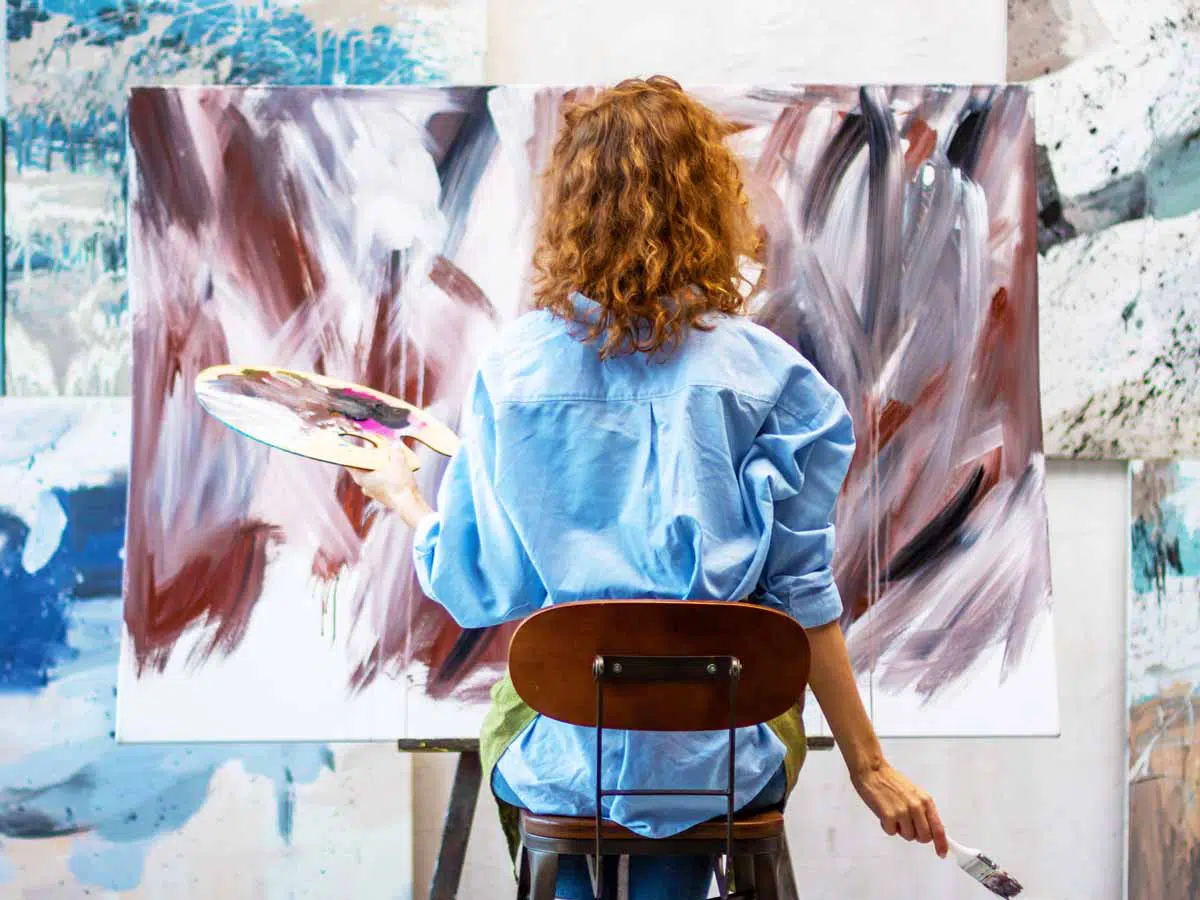 Femme de dos tenant une palette de couleur assise devant une oeuvre abstraite qu'elle peint.