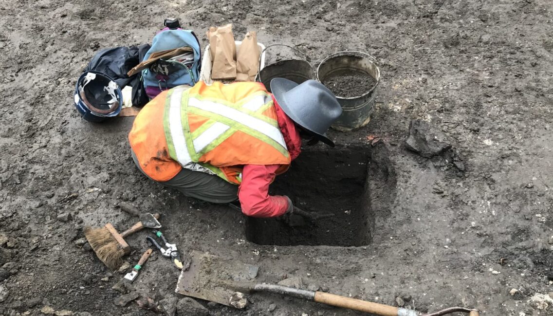 On voit une personne creuser le sol et effectuer une fouille archéologique.