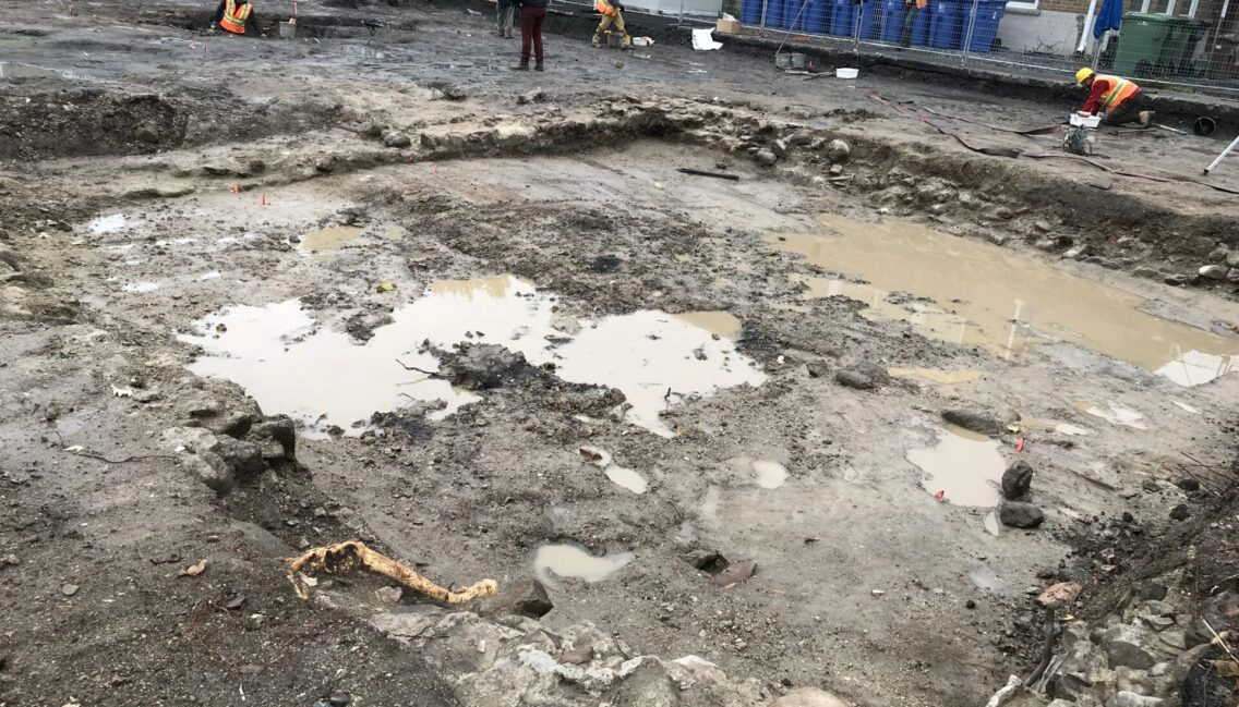 On voit plusieurs personnes travailler au sol. Elles effectuent des fouilles archéologiques sur un sol mouillé.